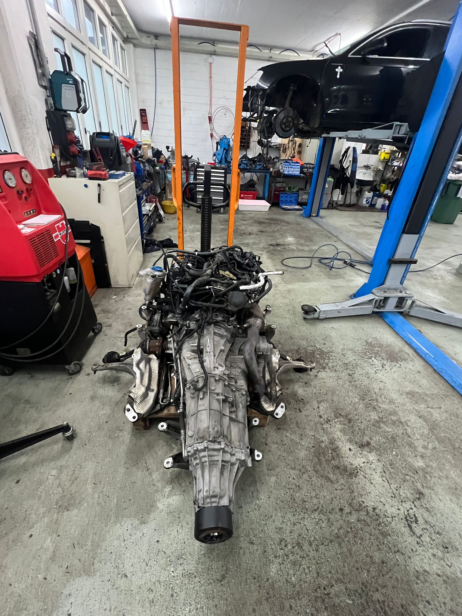 Ausgebauter Motor mit Getriebe am Boden der Werkstatt, dahinter ein Fahrzeug auf der Hebebühne
